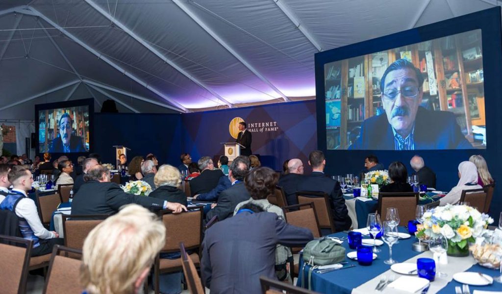 people at a ceremony listening to a person on a video screen speaking